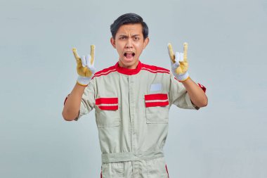 Yakışıklı genç tamircinin portresi çılgın bir ifadeyle elleri gri arkaplanda rock sembolü yapıyor.