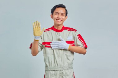 Mekanik üniforma giyen yakışıklı bir adamın portresi. Elleri göğsünde, avuçları açık gri arka planda küfrediyor.