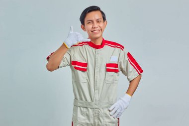 Mekanik üniforma giyen neşeli yakışıklı bir adamın portresi. Gri arka planda baş parmak işareti var.