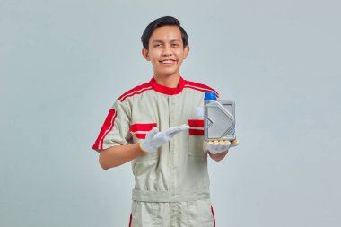 Motor yağı şişesini gri arka planda gösteren mekanik üniforma giyen gülümseyen yakışıklı adamın portresi.