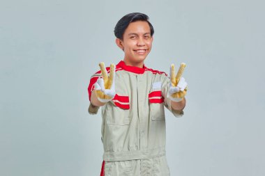 Tamirci üniforması giyen neşeli yakışıklı bir adamın portresi. Gri arka planda parmakları olan barış işareti yapıyor.