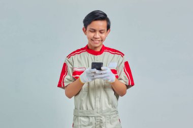 Gri arka planda izole edilmiş cep telefonu kullanarak mekanik üniforma giyen gülümseyen yakışıklı adam.