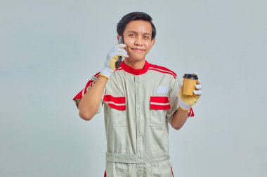 Akıllı telefondan konuşan ve elinde gri arka planda kahve tutan genç bir tamirci.