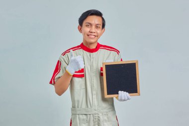 Boş tahta tutan yakışıklı genç tamircinin portresi ve gri arka planda gülümseyen bir ifadeyle baş parmak hareketi