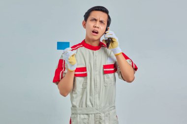 Şok olmuş yakışıklı tamirci akıllı telefondan konuşuyor ve gri arka planda kredi kartı tutuyor.