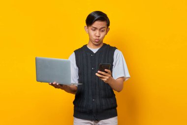 Üzgün yakışıklı genç adam cep telefonu ekranına bakıyor ve sarı arka planda dizüstü bilgisayarı tutuyor.