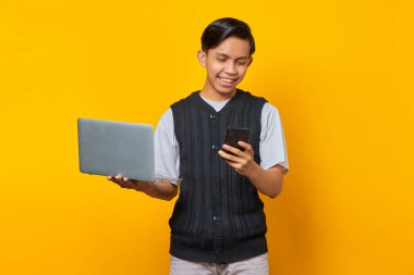 Neşeli yakışıklı genç adam cep telefonu ekranına bakıyor ve sarı arka planda dizüstü bilgisayarı tutuyor.