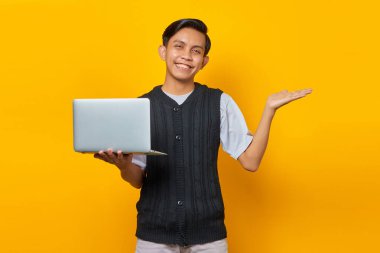 Gülümseyen yakışıklı genç adam avuç içinde kopya alanı gösteriyor ve sarı arka planda dizüstü bilgisayarı tutuyor.