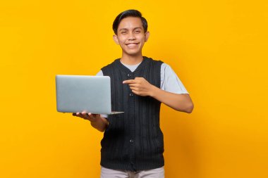 Gülümseyen yakışıklı genç adam sarı arka planda dizüstü bilgisayarı işaret ediyor.