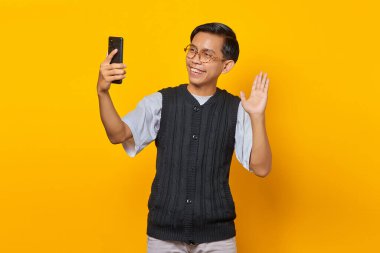 Cep telefonu tutan ve sarı arka planda el sallayan neşeli Asyalı genç adamın portresi.
