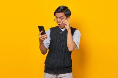 Şok olmuş Asyalı bir adam gözlüğe dokunuyor ve bir elinde cep telefonu, sarı arka planda.