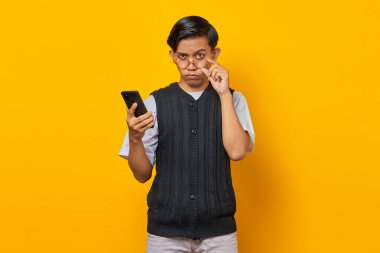Üzgün Asyalı adam gözlüğe dokunuyor ve bir elinde cep telefonu, sarı arka planda.