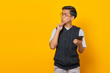 Sarı arka planda parmakları çenesinde cep telefonu kullanan düşünceli genç bir Asyalı.