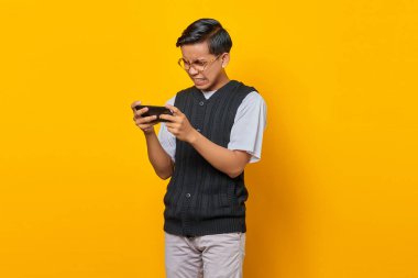 Sinirli Asyalı genç adam sarı arka planda cep telefonu oyunu oynuyor