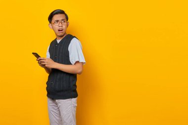 Şok olmuş genç ve yakışıklı bir adam akıllı telefon tutuyor ve sarı arka plana bakıyor.