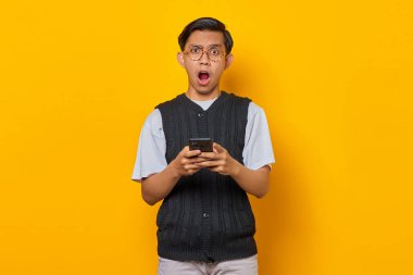 Sarı arka plan üzerinde cep telefonu kullanan, ağzı açık Asyalı genç bir adamın portresi.