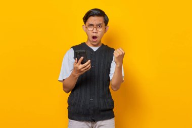 Sarı arka planda el hareketi başarısı olan mutlu Asyalı genç adam cep telefonu kullanıyor.