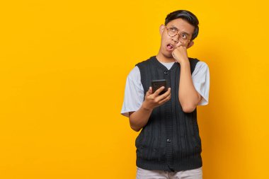 Eli çenesinde, akıllı telefonu sarı arka planda tutan dalgın Asyalı genç adamın portresi.