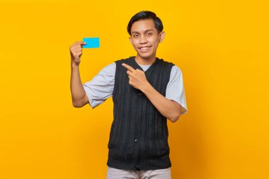 Neşeli genç Asyalı adam sarı arka planda kredi kartı gösteriyor.