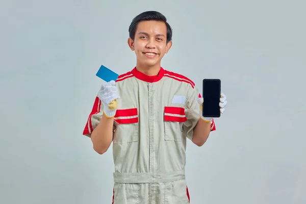 笑顔の若い整備士の肖像クレジットカードを示し 灰色の背景にスマートフォンを保持 — ストック写真