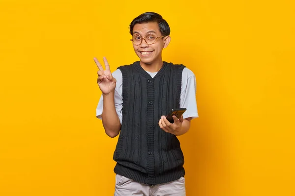 Веселий Молодий Азіат Тримає Мобільний Телефон Показує Знак Миру Жовтому — стокове фото