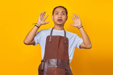 Şok olmuş ve şaşırmış Asyalı önlük giymiş genç bir adam ağzı açık ve iki elini de kaldırmış bir şekilde ileriye bakıyor.