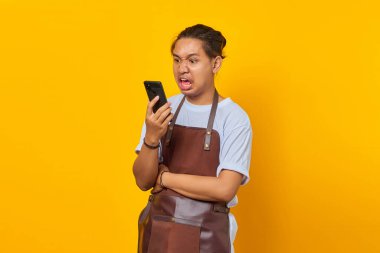 Önlük giymiş Asyalı bir gencin portresi. Sarı arka planda akıllı telefondan konuşurken sinirli görünüyor.