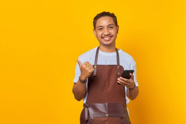 Cep telefonu tutan ve sarı arkaplanda izole edilmiş fotokopi alanını gösteren neşeli Asyalı genç adam.