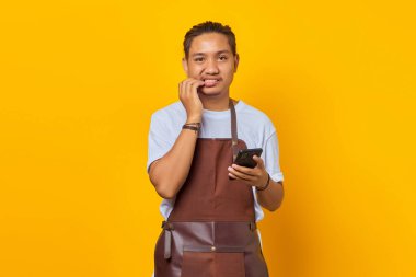 Akıllı telefon tutan Asyalı genç adam endişeli ve endişeli görünüyor. Sarı arka planda izole edilmiş bir şeyden korkuyor.