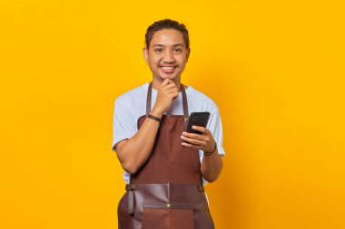 Sarı arka planda elleri çenesinde cep telefonu tutan önlük giymiş gülümseyen Asyalı genç adamın portresi.