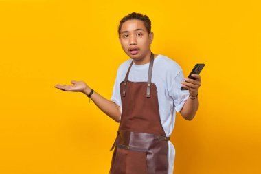 Sarı arka planda kafası karışık ve mutsuz bir ifadeyle akıllı telefon tutan önlüklü yakışıklı Asyalı genç adamın portresi.