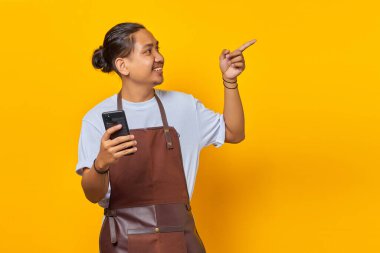 Önlük takan, akıllı telefon tutan ve sarı arka planda parmağını gösteren neşeli, yakışıklı Asyalı genç adam portresi.