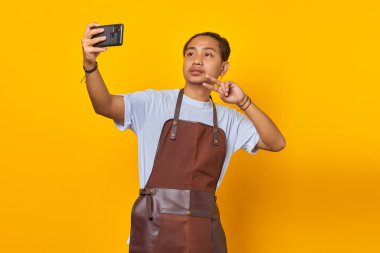Asyalı adamın portresi şaşırdı ve sarı arka planda cep telefonundan selfie çekerken barış işareti gösterdi.