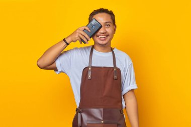 Sarı arka planda izole edilmiş akıllı telefonlu, önlük giyen yakışıklı Asyalı genç adamın portresi.