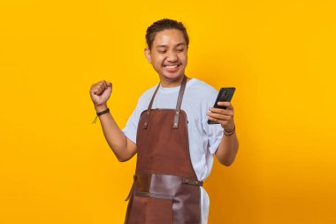 Önlük takmış, akıllı telefon tutan ve başarıyı el kaldırarak kutlayan heyecanlı Asyalı genç adamın portresi.