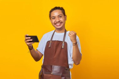 Cep telefonunda video oyunu oynayan ve sarı arka planda başarıyı kutlayan neşeli Asyalı genç adamın portresi