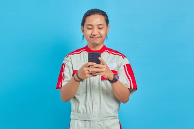 Mekanik üniforma giyen gülümseyen yakışıklı adam mavi arka planda cep telefonu kullanıyor.