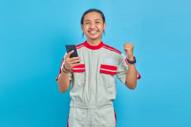 Mekanik üniforma giyen, akıllı telefon kullanan ve başarıyı mavi arka planda kutlayan neşeli yakışıklı adamın portresi.