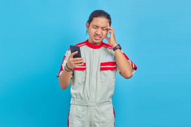 Mekanik üniforma giyen, akıllı telefon kullanan ve mavi arka planda baş ağrısı çeken yakışıklı bir adamın portresi