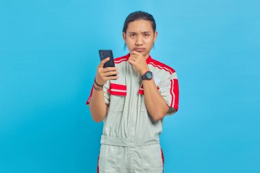 Mekanik üniforma giyen, akıllı telefon kullanan ve mavi arka planda kameraya bakan düşünceli yakışıklı adamın portresi.