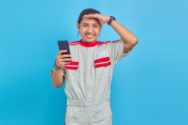 Mekanik üniforma giyen gülümseyen yakışıklı bir adamın portresi. Elinde akıllı telefon, mavi arka planda.