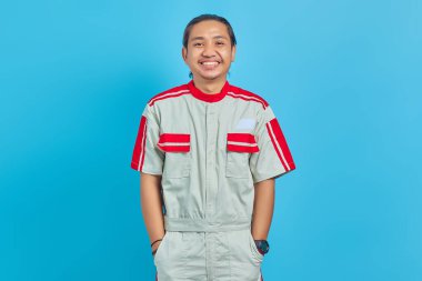 Üniformalı neşeli Asyalı tamirci mavi arka plandaki kameraya bakıp gülümsüyor.