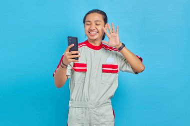 Mekanik üniforma giymiş gülümseyen yakışıklı bir adamın portresi video görüşmesi yapıyor ve mavi arka planda akıllı telefon kamerasına el sallıyor.