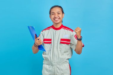 Elinde not panosu olan ve başarıyı mavi arka planda kutlayan neşeli, genç, yakışıklı mekanik bir adamın portresi.