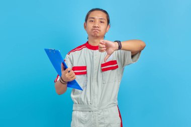 Mekanik üniforma giyen, elinde pano tutan ve mavi arka planda baş parmakları aşağıda işaretlerden hoşlanmayan sinirli yakışıklı bir adam.