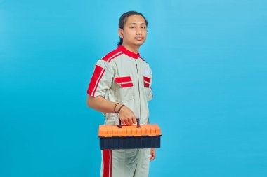 Mekanik üniforma giyen gülümseyen yakışıklı adamın portresi mavi arka planda izole edilmiş alet kutusunu tutuyor.