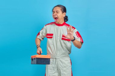 Mekanik üniforma giyen yakışıklı bir adamın portresi. Uykulu görünüyor ve mavi arka planda izole bir alet çantası tutuyor.