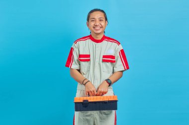 Mekanik üniforma giyen ve alet kutusunu mavi arka planda tutan neşeli yakışıklı adam.