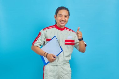 Mekanik üniforma giyen neşeli yakışıklı bir adamın portresi. Elinde pano ve mavi arka planda baş parmaklar var.