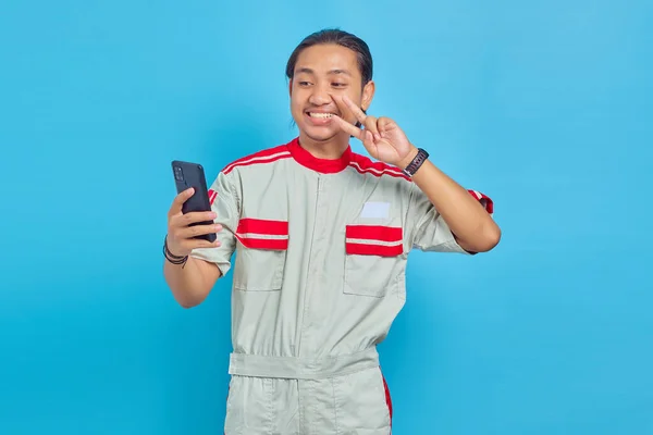 機械式制服を着た笑顔のハンサムな男の肖像ビデオ通話を作り 青の背景にスマートフォンのカメラに平和的なジェスチャーを示す — ストック写真